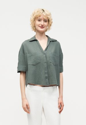 Νεαρή γυναίκα με σγουρά ξανθά μαλλιά, που φοράει ένα φαρδύ πράσινο πουκάμισο με κουμπιά και άσπρο παντελόνι, στέκεται μπροστά σε ένα απλό φωτεινό φόντο.