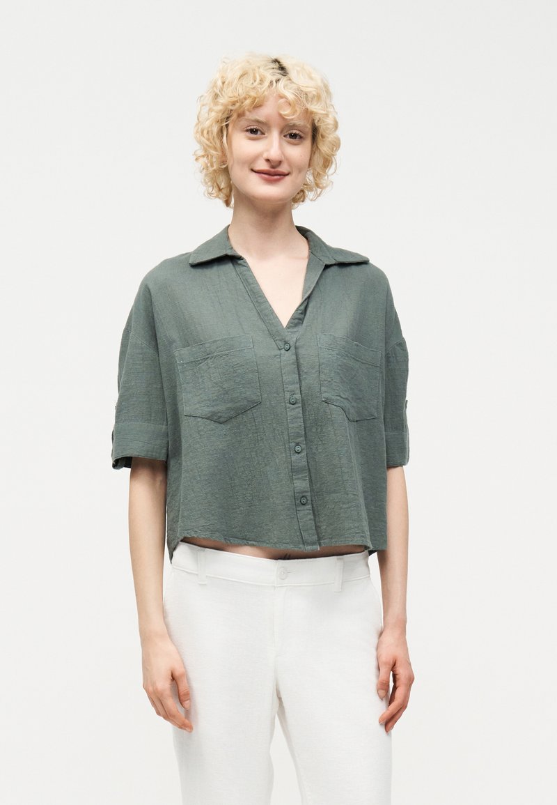 Νεαρή γυναίκα με σγουρά ξανθά μαλλιά, που φοράει ένα φαρδύ πράσινο πουκάμισο με κουμπιά και άσπρο παντελόνι, στέκεται μπροστά σε ένα απλό φωτεινό φόντο.