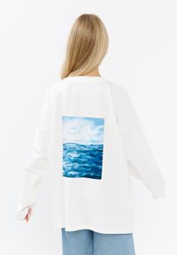 Biała koszula z długim rękawem z prostokątnym nadrukiem niebieskiej fali oceanicznej na plecach. Miękki bawełniany materiał, luźny krój, okrągły dekolt.