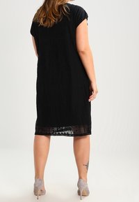 Zwarte jurk tot de knie met korte opgerolde mouwen, textuurstof, doorzichtige onderzoom, gecombineerd met lichtgrijze hoge hakken.