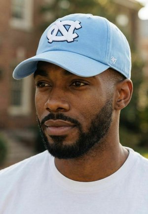 Mann mit Bart, der eine hellblaue UNC-Baseballkappe und ein weißes Hemd trägt, draußen mit unscharfem Backsteingebäude im Hintergrund.