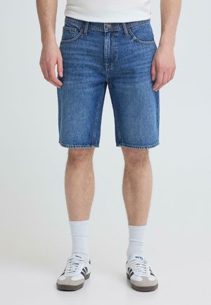 Mężczyzna w niebieskich jeansowych szortach, białych skarpetkach i białych trampkach z czarnymi paskami, stojący na prostym jasnym tle.