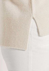 Beige gestrickter Pullover mit geripptem Textur, seitlichem Schlitz und langen Ärmeln, kombiniert mit weißen Hosen. Nahaufnahme des Stoffs und der Designdetails.