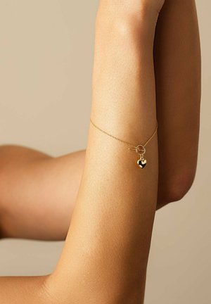 Goldarmband mit einer dünnen Kette, ausgestattet mit einem kleinen Herzanhänger mit einem schwarzen Akzent, getragen an einem nackten Arm vor einem neutralen Hintergrund.