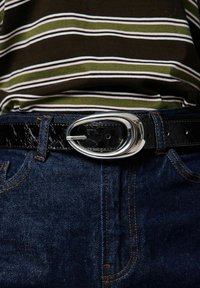 Juodas odinis diržas su blizgiu sidabriniu ovaliu sagtimi, dėvimas per tamsias džinsines kelnes, derinamas su dryžuota žalia ir rudai dryžuota palaidine.