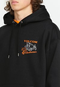 Czarna bluza z kapturem ze sznurkiem, z pomarańczowym i białym graficznym logo, które zawiera wizerunek ciężarówki oraz napis "VOLCOM Workwear". Miękka tekstura.