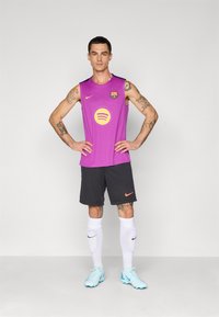 Tetoválásokkal rendelkező férfi lila FC Barcelona ujjatlan mezben, fekete rövidnadrágban, fehér zokniban és világoskék cipőben áll, kezét a csípőjén támasztva.