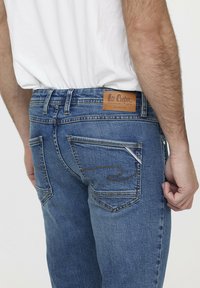 Jean en denim bleu avec un lavage moyen, présentant une étiquette en cuir, des poches cousues et une coupe droite. Vue arrière simple.