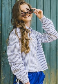 Jonge vrouw met lang krullend haar die glimlacht terwijl ze een zwarte zonnebril optilt, gekleed in een gestreept shirt en blauwe broek tegen een groene houten muur.