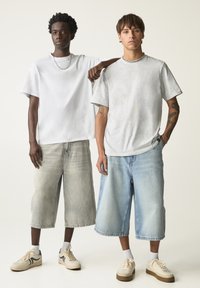 Dva modelky mají na sobě oversized trička, jedno bílé a jedno světle šedé, kombinované s džínovými šortkami ke kolenům. Obě mají na sobě tenisky s bílými detaily.