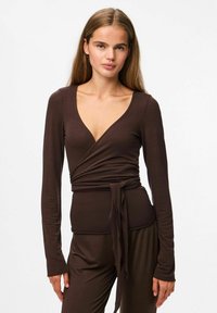 Jauna sieviete ar gariem matiem, ģērbta cieši pieguļošā brūnā apģērbā ar dziļu V veida kaklu un atbilstošajās brīvās brūnās biksēs, pret vienkāršu baltu fona.