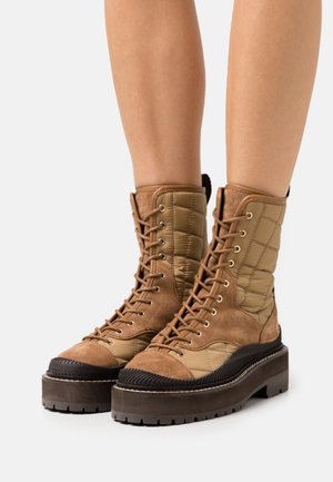 Bottes marron matelassées à lacets avec semelles épaisses portées sur des jambes nues sur un fond clair uni.