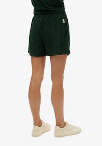 Pantalones cortos de color verde oscuro hechos de una tela texturizada con un patrón. Cintura elástica y bolsillos laterales. Llevados con zapatillas de color claro.