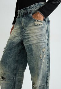 Hellblaue Jeans aus Denim mit einem ausgebleichten Look, die abgenutzte Details, große Risse und einen geraden Schnitt aufweisen. Zwei sichtbare Seitentaschen.