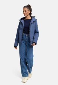 Mėlynas vandeniui atsparus džemperis su kapišonu, priekiniu užtrauktuku ir kontrastuojančia juoda išklotine, dėvimas ant tamsiai mėlynos palaidinės ir šviesiai mėlynų džinsų, kartu su baltais batais.