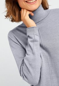 Pull en col roulé gris avec une texture douce. Il présente une coupe ajustée, des poignets côtelés et une apparence sans couture au niveau des épaules et des manches.
