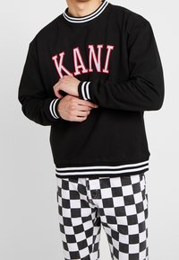 Man wearing black sweatshirt with "KANI" in red letters and black-and-white checkered pants, hands clasped at waist.