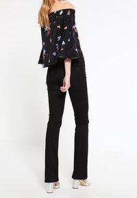 Top nero a fiori con spalle scoperte e maniche svasate, abbinato a jeans neri aderenti e scarpe con tacco argentate, visto da dietro.