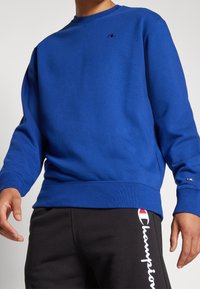 Blå sweatshirt med rund hals, ribbade muddar och nederkant. Har en liten logotyp på bröstet och en strukturerad tyg. I kombination med svarta sweatpants.