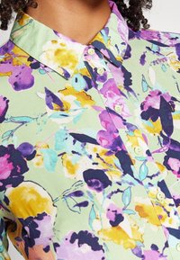 Vihreä kukallinen nappipaitapusero, jossa on purppuran, keltaisen ja pinkin kuvioita; puuvillakangasta; leveä kaulus ja vaaleanvihreät napit.