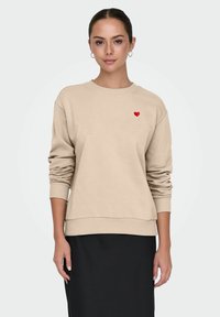 Jeune femme portant un sweat-shirt beige avec un petit cœur rouge sur la poitrine et une jupe noire, debout devant un fond clair uni.