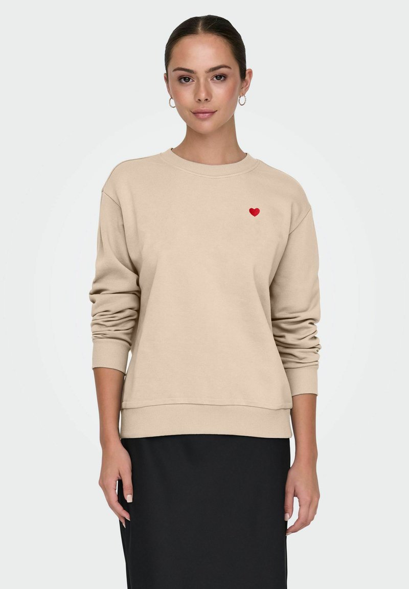 Jeune femme portant un sweat-shirt beige avec un petit cœur rouge sur la poitrine et une jupe noire, debout devant un fond clair uni.