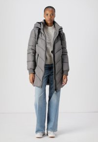 Doudoune grise avec rembourrage en chevrons, fermeture éclair à l'avant et capuche. Portée par-dessus un pull gris clair et assortie à un jean bleu clair et des baskets blanches.