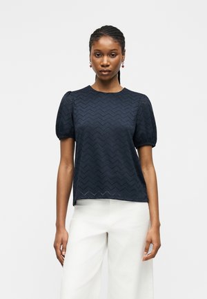 Femme portant un chemisier bleu marine à manches courtes avec un motif œillet en zigzag et un pantalon blanc à jambes larges, debout devant un fond uni.