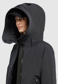 Giacca nera con un ampio cappuccio oversize, chiusura con zip e tasche laterali. Il tessuto appare liscio con finitura opaca e dettagli minimi.