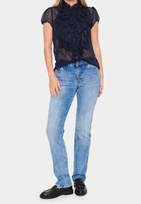 Donna che indossa una camicetta trasparente blu navy con volant, jeans dritti azzurri chiari e mocassini neri in pelle, in piedi contro uno sfondo neutro.