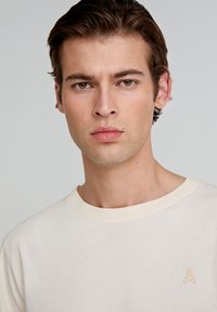 Camiseta de algodón crema con cuello redondo y mangas cortas, que presenta un pequeño diseño bordado de calavera y tibias cruzadas en el pecho.