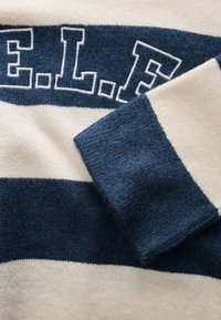 Jersey de punto a rayas azul marino y beige con letras en negrita azul marino delineadas en blanco "E.L.F." en el pecho y una manga doblada.