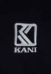 Tissu noir avec un logo brodé comportant des formes géométriques blanches, y compris des lettres et une bordure rectangulaire.