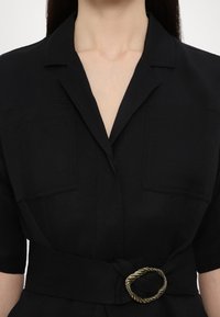 Rochie neagră cu brațel, având guler de cămașă, două buzunare pe piept, mâneci scurte și un accent cu cataramă în stil răsucit. Fabricată dintr-un material texturat.