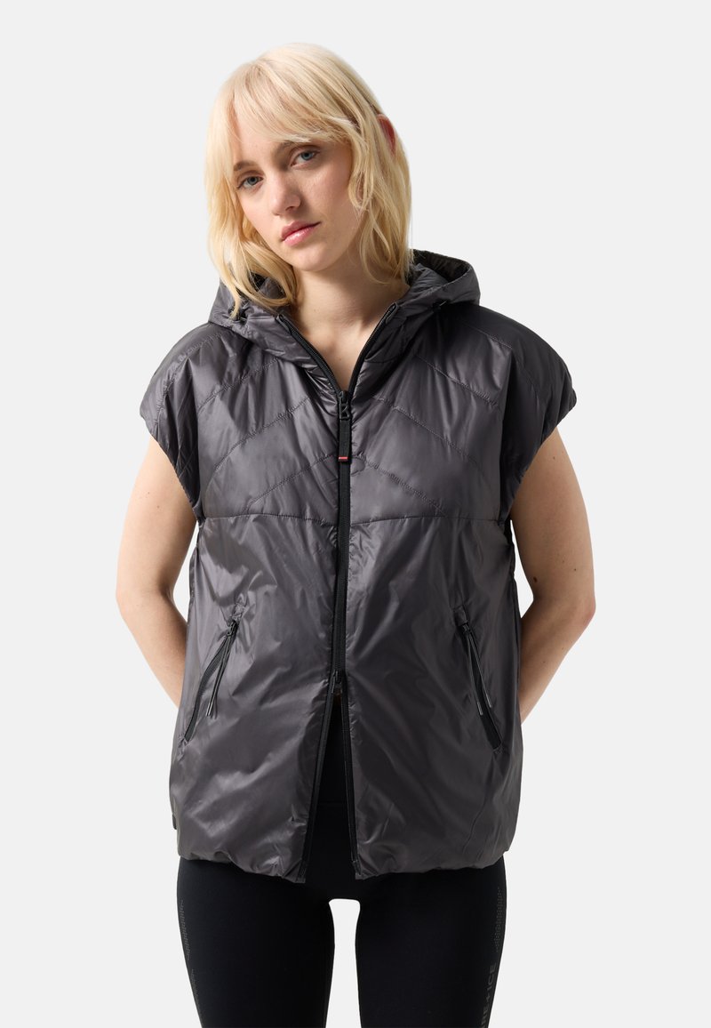 Lyshåret kvinde iført en ærmeløs, mørkegrå hætte-puffervest med lynlåslommer, stående mod en lys grå baggrund.