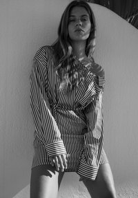 Schwarz-weißes Bild eines Models, das ein locker sitzendes, gestreiftes Hemd mit einem schulterfreien Ärmel und Knopfdetails trägt.