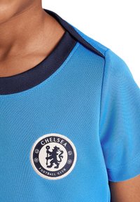 Maglia da calcio blu con maniche corte, caratterizzata da un distintivo rotondo del Chelsea Football Club in bianco e blu navy sul petto. Materiale in misto cotone.