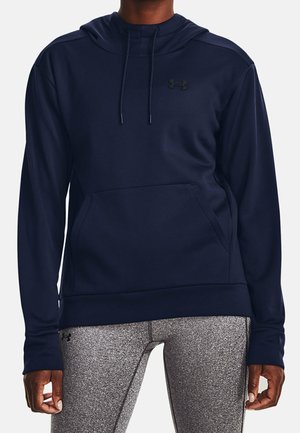 Persoon draagt een donkerblauwe capuchonsweater met een voorzak en grijze gestructureerde leggings, staat met ontspannen armen langs het lichaam.