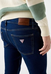 Ciemnoniebieskie dżinsy z denimu z skórzaną naszywką na pasku i trójkątnym czerwono-białym logo na tylnej kieszeni.