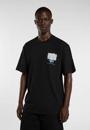 Uomo dalla pelle scura che indossa una t-shirt nera oversize con logo e grafica "Dickies" blu e bianca sul petto sinistro, in piedi davanti a uno sfondo semplice.
