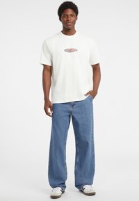 Bílý tričko s krátkým rukávem s logem "GUESS JEANS", zkombinované se volnými světle modrými džínami a bílými teniskami. Čisté pozadí.