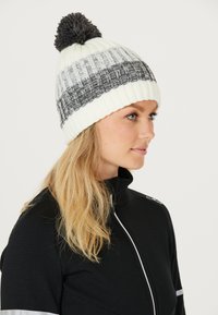 Vit och grå stickad mössa med ett texturerat mönster och en grå pom-pom på toppen, med en ribbad kant för en snug passform.