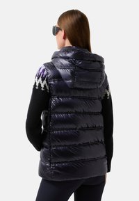 Frau, die eine schwarze Pufferweste mit Kapuze über einem schwarzen Pullover mit lila und weißen Mustern trägt, mit dem Rücken zu uns gewandt, langen braunen Haaren und einer schwarzen Sonnenbrille.