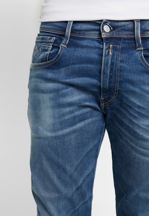Gros plan d'un jean en denim bleu porté avec un haut blanc, montrant les poches avant, les passants de ceinture et la fermeture par bouton.