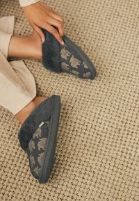 Dark gray slippers with a fluffy gray trim, featuring a patterned upper with gold bears. Non-slip sole and textured surface. Woven rug background.
