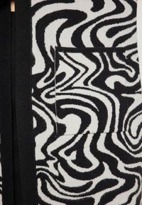 Fekete-fehér mintázatú textil, forgó vonalakkal, látható zsebfelnakítéssel, texturált gyapjúkeverék anyag, közeli nézet.