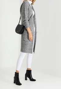 Długi szary kardigan z dzianiny z kapturem, kieszeniami z przodu, noszony na białym golfie, zestawiony z czarnymi lakierowanymi botkami za kostkę i torebką na ramię.