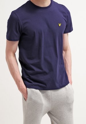 Mężczyzna ubrany w jednolity ciemnoniebieski krótki rękaw z małym żółtym logo na piersi i jasnoszare spodnie, z rękami w kieszeniach.