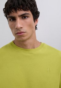 Un jeune homme aux cheveux noirs bouclés portant un t-shirt col rond vert citron avec un petit crâne et des os croisés brodés sur la poitrine.