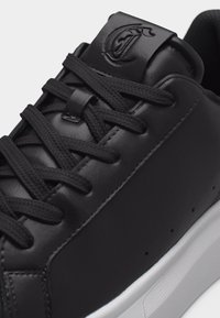 Κοντινή λήψη μαύρου δερμάτινου αθλητικού παπουτσιού με μαύρα κορδόνια, αφρώδες γλώσσα με ανάγλυφο λογότυπο και αντίθετη λευκή σόλα.